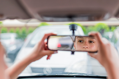 Teen girl adjusting rearview mirror during the day while driving her new, first  car. Focus on girl in the mirror.