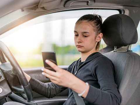 Young woman or female teen looking to her smartphone while driving car during bright suuny day - side view, sun shines through front window. Concept of danger driving.