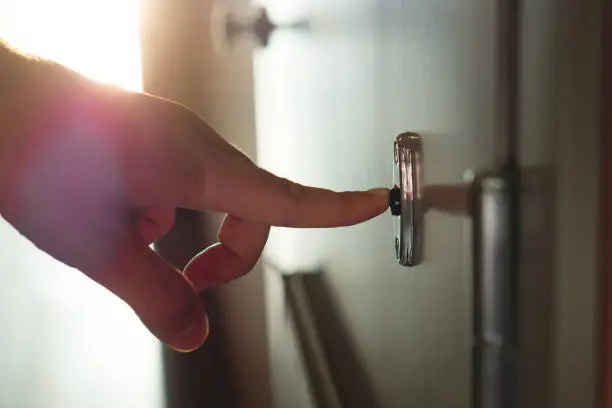 Photo of Finger pressing doorbell in sunny apartment building corridor. Close up of male hand ringing door bell in a block of flats. Salesman, fundraiser, guest or visitor behind door.