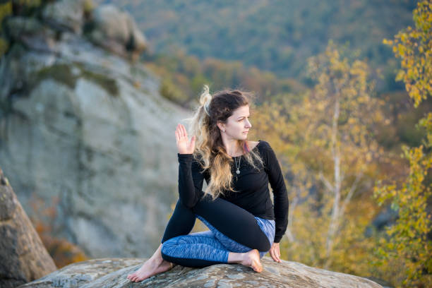 Sporty fit woman is practicing yoga on the top of the mountain Calm young woman is practicing yoga and doing asana Arha Matsyendrasana on the top of the high rocky mountain in the evening. Autumn forests, rocks and hills on the background arhanes stock pictures, royalty-free photos & images