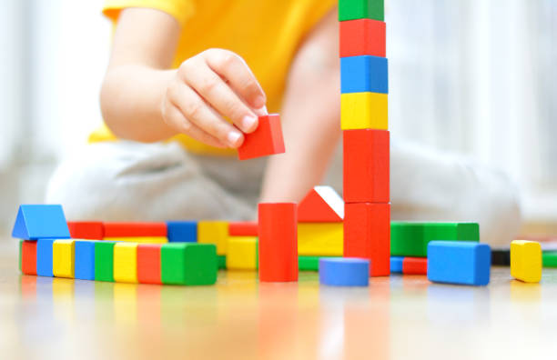 A five year old boy in yellow t-shirt is playing on the parquet floor. He is made tower of cubes.