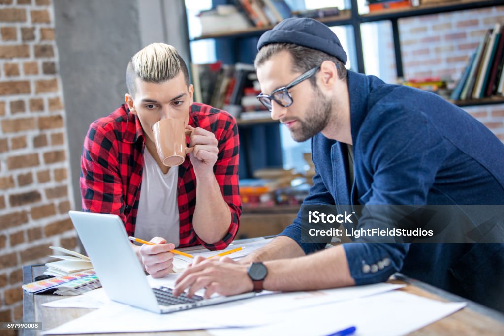 Designers working at project Two young male designers working at project while using laptop and drinking coffee Adult Stock Photo