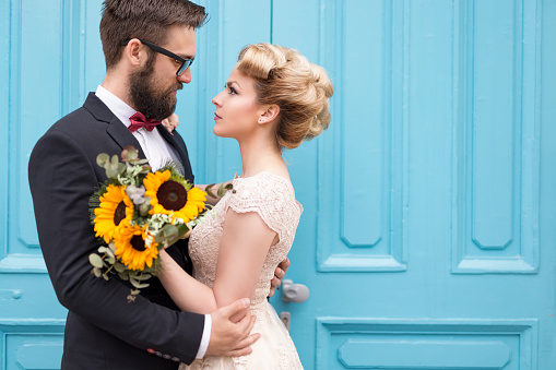 Newlywed couple standing next to a blue retro wooden door, holding a sunflower wedding  bouquet and looking at each other