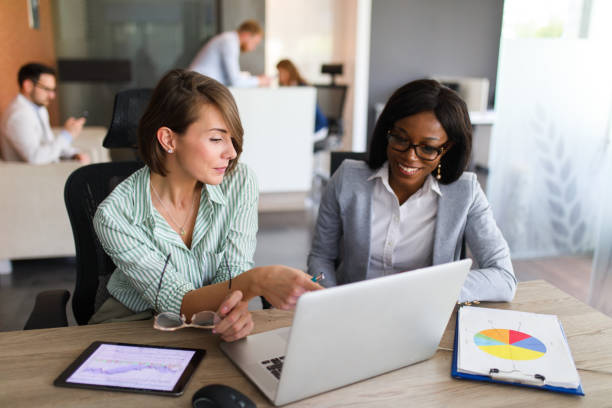 Businesswomen with laptop, digital tablet and chart papers cooperating Two businesswomen are in the office using laptop, digital tablet and chart papers while working together. progress report stock pictures, royalty-free photos & images