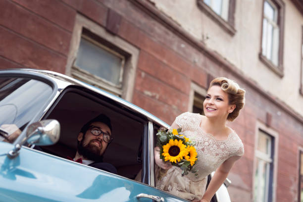 We do Young newlywed couple in a retro vintage car, groom driving while bride is waving through a window while they are leaving on their honeymoon. Focus on the bride vintage car photos stock pictures, royalty-free photos & images