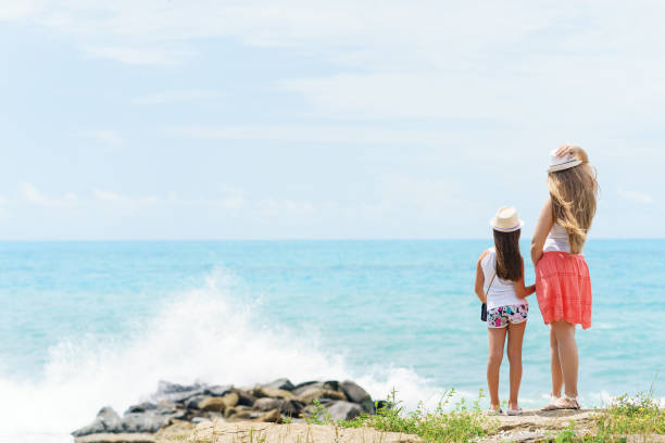 young european woman and her little daughter or younger sister are staying and holding hands of each other at the shore of blue sea with strong splaching waves - splaching imagens e fotografias de stock