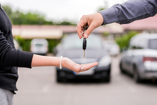transportation and ownership concept - customer and salesman with car key outside. Woman at a car dealership buying an auto, the sales rep giving her the key, macro shot with focus on hands and key