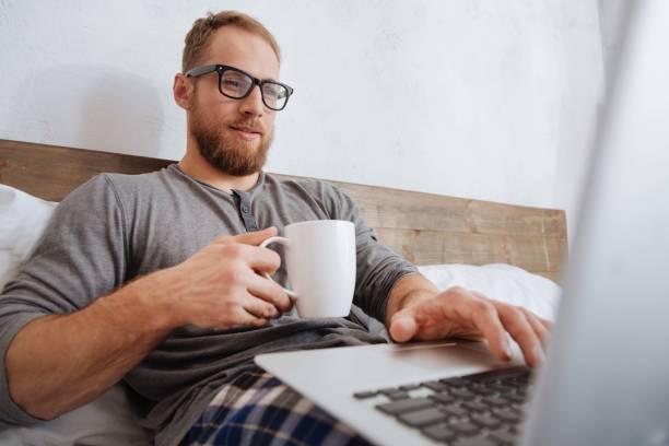 istock Cheerful man drinking coffee and working with laptop in bed 698026172