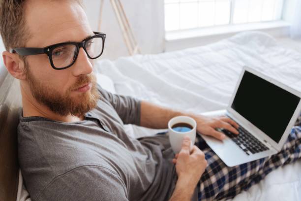istock Bearded man sitting on bed with coffee and laptop 698026244