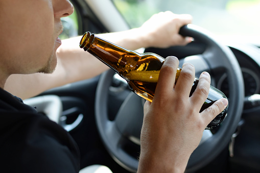 The concept of alcohol driving crime - closeup of young male driver hands with steering wheel and bottle of beer.