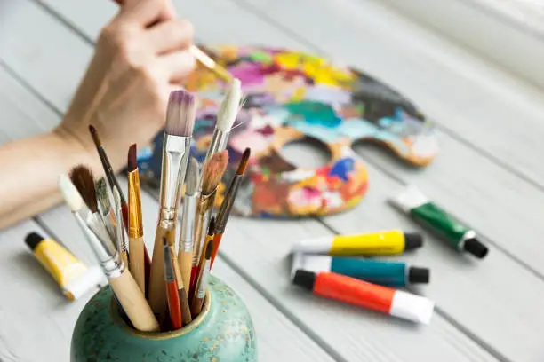 A variety of paint brushes in the foreground,Hands holding a brush and a pallet for mixing paints with tubes of acrylic paint in the background.