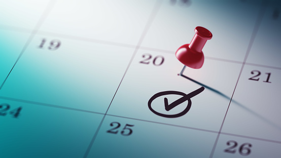 Concept image of a Calendar with a red push pin. Closeup shot of a thumbtack attached. Check Mark written on a white notebook to remind you an important appointment.