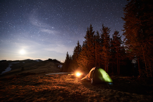 Tourist camping near forest in the night. Illuminated tent and campfire under beautiful night sky full of stars and the moon