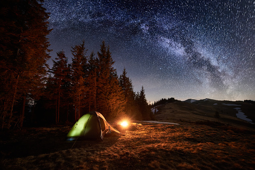 Night camping. Illuminated tent and campfire near forest under beautiful night sky full of stars and milky way