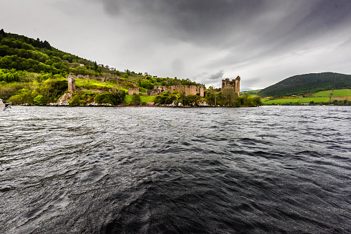 Ruins of the beautiful Urquhart castle and the hills containing the castle taken during boat ride on the loch ness. The gloomy day and the clouds provide a dramatic effect to the ruins.