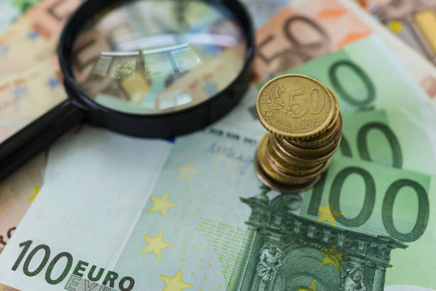 Stack of Euro coins on pile of banknotes with magnifying glass as business financial tax concept stock photo