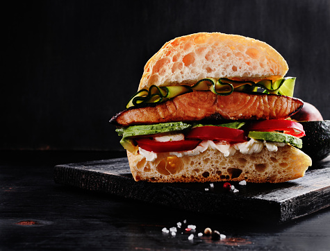 Ultimate sandwich with grilled salmon, cream cheese, sliced avocado and seasonals on black wooden board with free text space.