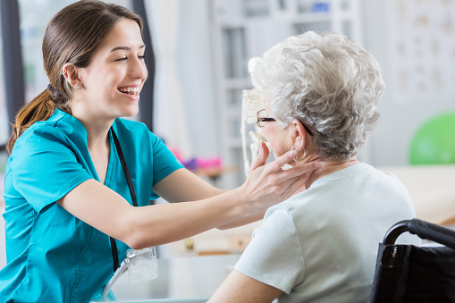 Attractive young nurse cracks a joke and laughs with her patient to ease tension as she palpates her neck muscles.  The wheelchair bound senior woman is complaining of severe neck pain.