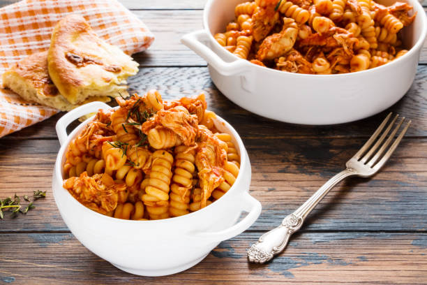 fusilli pasta with chicken. cooked in spicy sauce from tomatoes, onion, garlic, dried oregano and thyme, paprika and olive oil. fresh bread, white bowl and casserole on wooden brown rustic table. - oregano freshness herb brown imagens e fotografias de stock