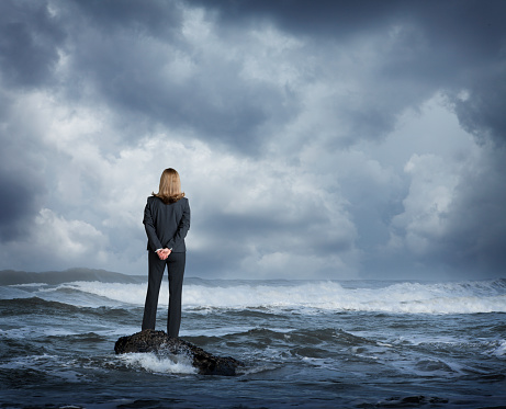 A rear view of a businesswoman as she stands on a small rock in a rough ocean surf.  She stands with her hands clasped behind her back as she looks out across the rough waters and out towards the stormy sea.