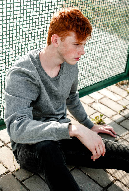 한 젊은 잘생긴 빨간 머리 유행 소년 야외, 화창한 날에 경기장 근처에 앉아 캐주얼 옷을 입고. 가까이, retouched 샷. - prepossessing 뉴스 사진 이미지