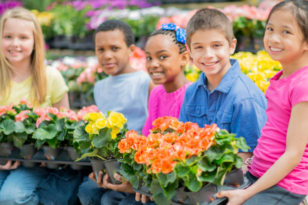 A multi-ethnic group of elementary age school children are on a field trip and holding colorful flowers inside pots in a greenhouse nursery. They are looking at the camera and smiling.
