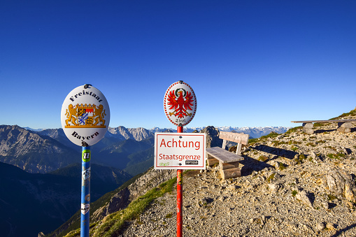Two sign posts at the border between the German state of Bavaria and the Austrian state of Tirol located on a mountain ridge in the Karwendel mountains in the Alps. Warning sign: \