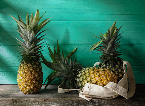 Market fresh tropical pineapple standing next to more pineapples in a reusable cotton bag on a rustic wooden table against a turquoise wooden walled background. Good copy space above the fruit.