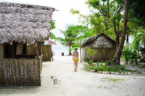Detail of Kuna Yala village with tourist cottages and senior woman walking on path. In front her is lokal market and some tourists on beach. It is on small  Island Naranjo Chico in San Blas archipelagos in Caribbean See, Panama.  Cottage is from wood, and bamboo.  San Blas is an old name for Panamas northeast province known for Kuna Yala indigenous people living there. Official they are Gunas and they live in Guna Yala province. The people are very small and mostly they don’t have connections with other people and cultures.