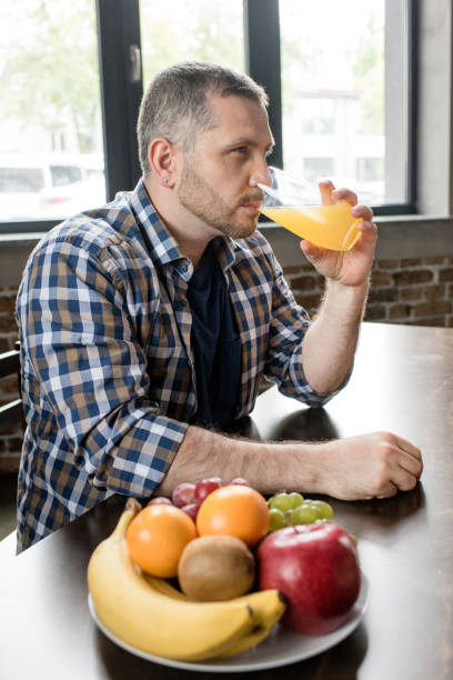 istock Bearded man drinking orange juice while sitting at kitchen table with fruits on plate 698620352