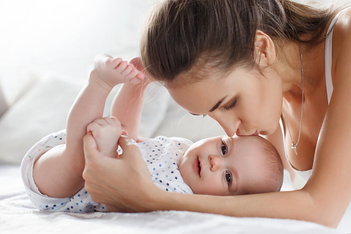 Beautiful young woman,brunette with thick hair braided in a thick braid, makeup and pleasant smile,dressed in blue trousers and a white jersey,playing with her newborn son in the bedroom on the bed,a boy with short,fluffy hair in a white T-shirt