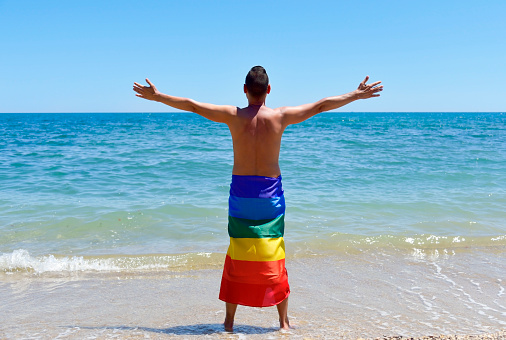 a young caucasian man seen from behind wrapped in a rainbow flag in front of the ocean