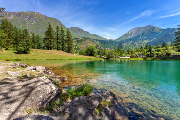 A hiker with a backpack and their dog traverse rocky terrain alongside a serene alpine lake, surrounded by a rugged mountain landscape. The scene evokes a sense of adventure and the beauty of nature in a remote, peaceful setting.