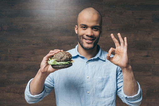 Handsome smiling young man holding tasty hamburger and showing ok sign