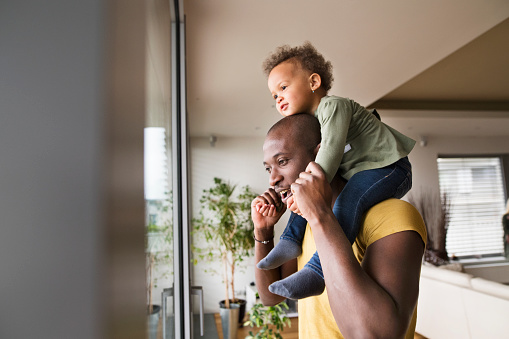 Young afro-american father at home at the window carrying his little daughter on his shoulders.