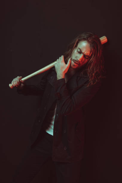 Confident young long haired man holding baseball bat and touching chin in studio