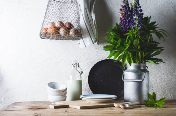 Photo of Concept of rural kitchen. Lupines in a can, milk, eggs and ware on a wooden table against the background of a white wall