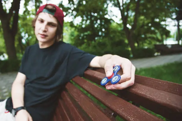 Photo of A long-haired hipster in a cap sits on a bench and spins a fidget-spinner