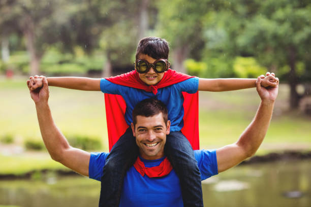 Boy in superhero costume sitting on fathers shoulders in park