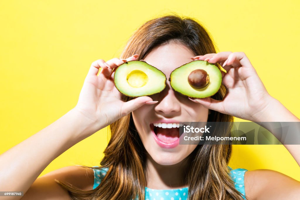 Happy young woman holding avocado halves Happy young woman holding avocado halves on a yellow background Avocado Stock Photo