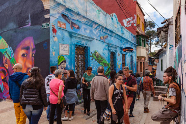 Bogotá, Colombia - Tourists and Local Colombian People on the Narrow Calle Del Embudo in the Historic La Candelaria District of the Andean Capital City Bogotá, Colombia - May 28, 2017: Tourists and local Colombian people seen on the colourful Calle del Embudo where it joins the Plaza del Chorro de Quevedo. The Street gets its name from its shape. Embudo translated into English is Funnel.  The Street is shaped like a funnel; this is the narrow end of the Street. It is so narrow that it is only accessible by foot.  The external walls of the buildings on the street are pained with street art that often depict the legends of the Pre-Colombian era. It is in this area that the Spanish Conquistador Gonzalo Jiménez de Quesada established the capitl city of Bogota in 1538. Photo shot in the afternoon sunlight; horizontal format. Camera: Canon EOS 5D MII. Lens: Canon EF 24-70 F2.8L USM. calle del embudo stock pictures, royalty-free photos & images