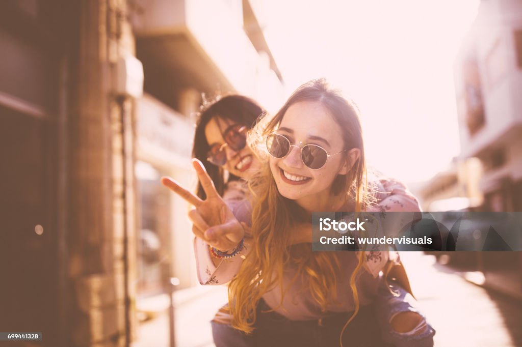 Young woman on piggyback ride doing the peace sign Hipster teenage girl on piggyback ride in the city doing the peace sign Friendship Stock Photo