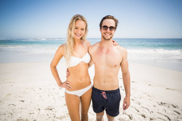 Portrait of young couple standing on the beach Portrait of young couple standing on the beach on a sunny day male swimsuit standing arm around stock pictures, royalty-free photos & images