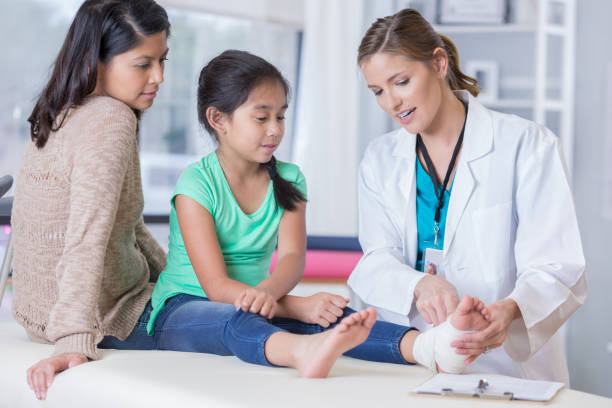 Confident emergency room doctor with young injured patient Emergency room doctor talks with young girl and the girl's mom about the girl's injured ankle. The doctor is pointing to the girl's wrapped ankle. emergency medicine stock pictures, royalty-free photos & images