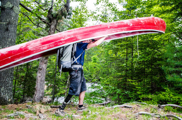 Man, seen from side, with prosthetic leg carrying his canoe through a forest footpath to the river  during a summer day