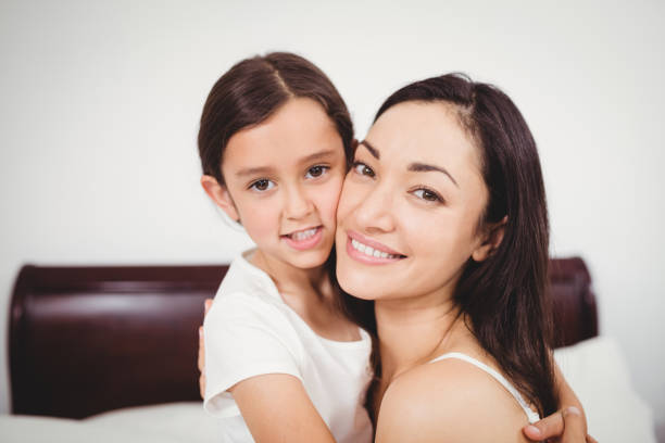 Close-up portrait of mother smiling while hugging daughter on bed at home