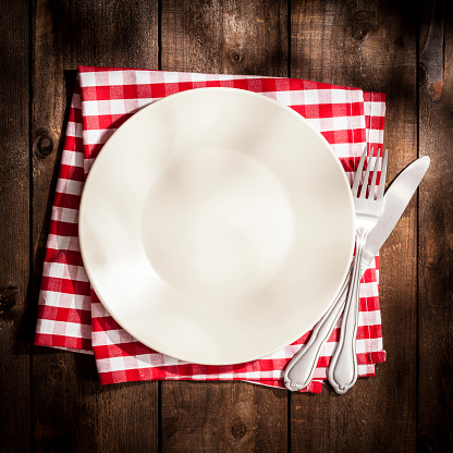 Top view of an empty plate on a red and white checkered textile napkin ready for menu text or any food with a fork and knife beside it shot on rustic wooden table. Predominant colors are brown and red. Low key DSRL studio photo taken with Canon EOS 5D Mk II and Canon EF 100mm f/2.8L Macro IS USM