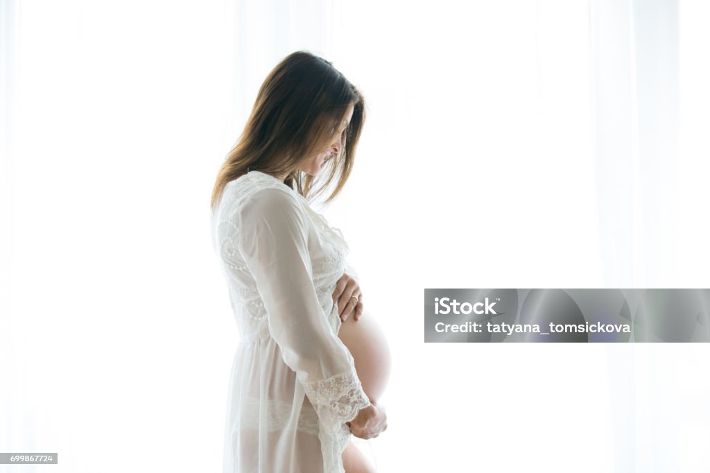 Portrait of young pregnant attractive woman, standing by the window, Portrait of young pregnant attractive woman, standing by the window, dressed in white dress, isolated image Abdomen Stock Photo