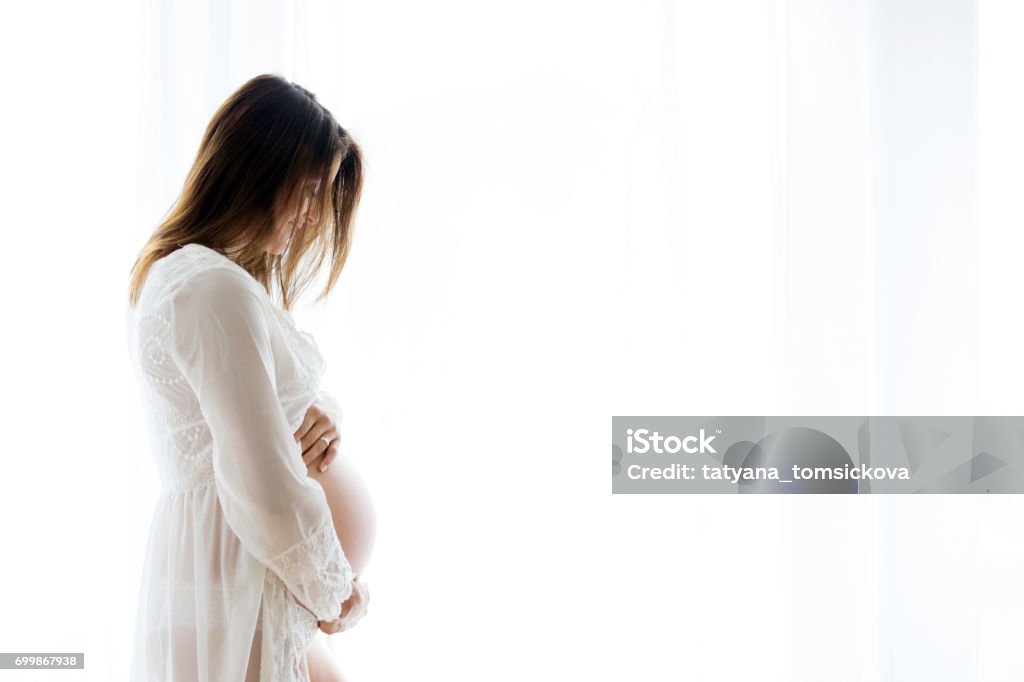 Portrait of young pregnant attractive woman, standing by the window, Portrait of young pregnant attractive woman, standing by the window, dressed in white dress, isolated image Abdomen Stock Photo