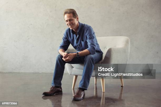 Casual Middle Aged Man Smiling While Sitting On Armchair Indoors Stock Photo - Download Image Now